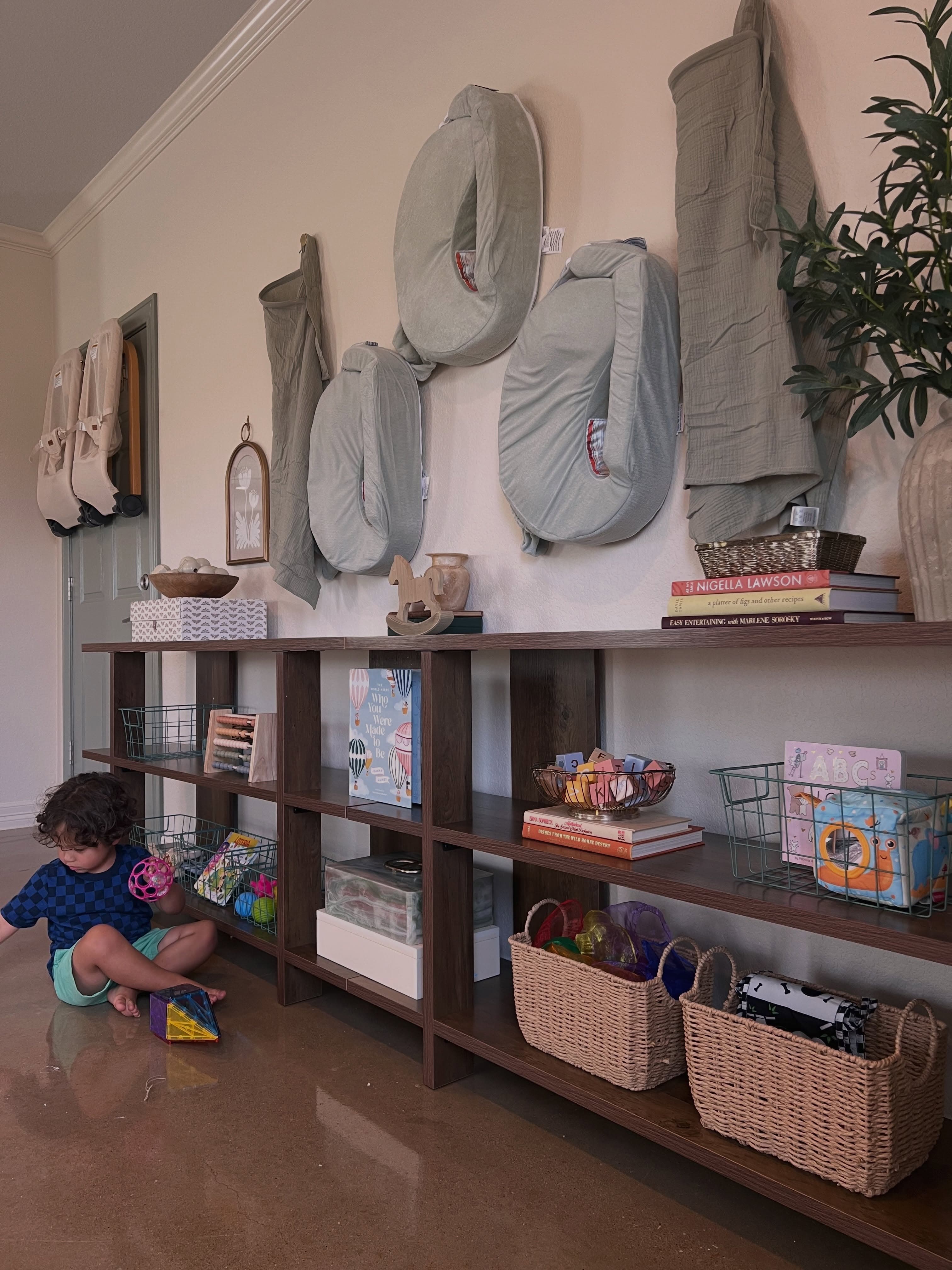 A child playing beside low shelves organized with toys, books, baskets, and feeding essentials